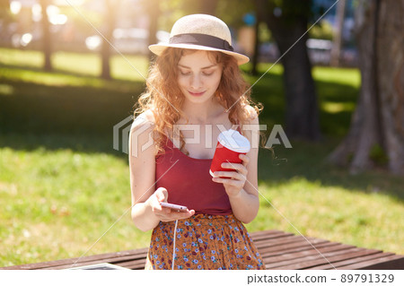 Image of woman with foxy hair sitting on bench in city park, charging mobile phone on bench via solar panel, holding takeaway coffee and checking her social network. Modern technology concept. 89791329