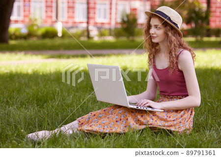 Freelance business concept. Woman sitting on green grass lawn in city park, working on laptop, has pensive facial expression, looks aside. Student girl studying outdoors, using wireless Internet. Freelance business concept. Woman sitting on green grass lawn in city park, working on laptop, has pensive facial expression, looks aside. Student girl studying outdoors, using wireless Internet. 89791361