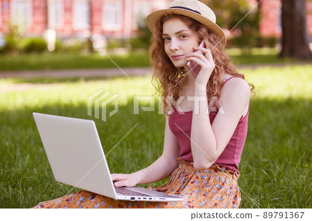 Shot of young woman in hat, sitting on green grass and using laptop for online education in park, has conversation with her teacher via smart phone, looking aside while comunicating, posing outdoor. Shot of young woman in hat, sitting on green grass and using laptop for online education in park, has conversation with her teacher via smart phone, looking aside while comunicating, posing outdoor. 89791367