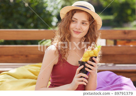 Summer and people concept. Young woman or teenage girl wearing casual clothes, holdinmg ice cream in hands, looking mysteriously aside, spending time with friends, posing over city park background. 89791379
