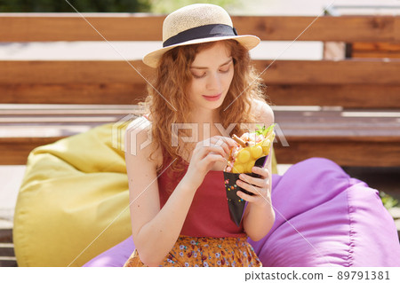 Image of sweet cute female having fair curly hair, looking attentively at ice cream, having meal, enjoying her weekends alone in park, sitting at colorful beanbag, wearing straw hat. Youth concept. 89791381