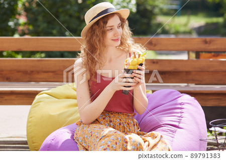 Outdoor shot of young female with foxy curly hair, sits on frameless chair smiling, looking aside and holding big ice cream in her hands, being in park in summer, cools with ice cream in hot weather. 89791383