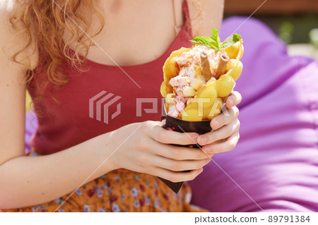Portrait of unknown woman holding big yellow ice cream while sitting on purple frameless chair, isolated on city park background, woman spending hot summer day in open air with her boy friend. 89791384