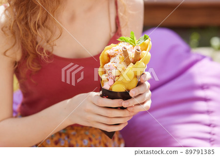Photo of slender lady with curly hair wearing red top, colorful skirt, sitting on violet beanbag outdoors alone, holding dessert in both hands, having street food, going to get rid of hunger. 89791385