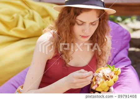 Outdoor shot of Caucasian charming woman eating healthy enjoying frozen treat snack in summer park, wearing casual clothes and hat, sitting on frameless mild chair, relaxing after exame in university. Outdoor shot of Caucasian charming woman eating healthy enjoying frozen treat snack in summer park, wearing casual clothes and hat, sitting on frameless mild chair, relaxing after exame in university. 89791408