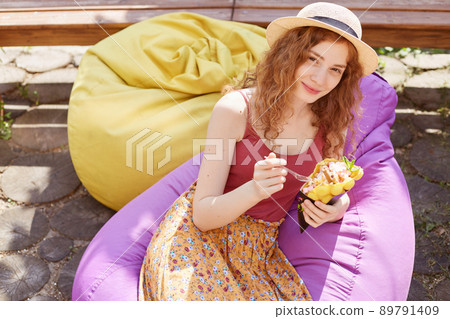 Outdoor shot of charming woman sitting on big purple cushioned frameless chair in town park, dressed casual burgunde t shirt, skirt and hat, eating ice cream, being photographed by her friend. 89791409