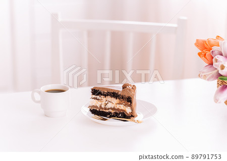 Piece of cake on white plate next to fork and cup of coffee on white table Piece of cake on white plate next to fork and cup of coffee on white table 89791753
