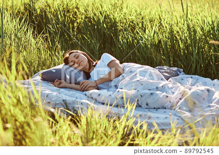 Young attractive dark haired female lying on soft pillow under blanket outdoor, keeps eyes closed, sleeping in open air, enjoying warm summer time, relaxing on nature. 89792545