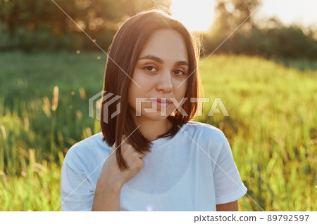 Cute calm female wearing white casual T-shirt posing outdoor in the middle of green field, looking directly at camera, looks relaxed and rested, woman in beautiful nature. 89792597