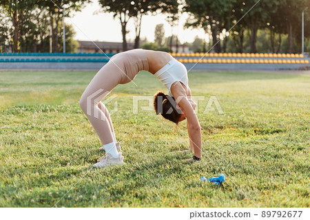Portrait of slim young adult woman backbending on stadium field, working out outdoor, training alone, health care, healthy lifestyle, sport activity in summer. 89792677