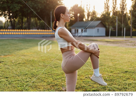 Side view portrait of brunette female wearing white top and beige leggins warming up before working out in the stadium, stretching leg, looking in distance. 89792679