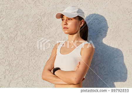 Portrait of serious confident female wearing white top and visor cap, standing isolated over gray wall outdoor with folded hands, looking in distance, sporty woman after work out. Portrait of serious confident female wearing white top and visor cap, standing isolated over gray wall outdoor with folded hands, looking in distance, sporty woman after work out. 89792833