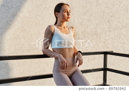 Young adult confident female with dark hair and ponytail wearing white top and beige leggins standing, looking away, relaxing after training, standing near handrail outdoor. Young adult confident female with dark hair and ponytail wearing white top and beige leggins standing, looking away, relaxing after training, standing near handrail outdoor. 89792842