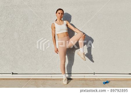 Attractive fitness model girl posing near gray wall wearing white sport top and beige leggins, standing on one leg, looking directly at camera with confident expression. Attractive fitness model girl posing near gray wall wearing white sport top and beige leggins, standing on one leg, looking directly at camera with confident expression. 89792895