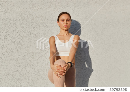 Outdoor shot of young of young age wearing white top, standing on one leg, touching knee with hands, looking at camera with serious facial expression. Outdoor shot of young of young age wearing white top, standing on one leg, touching knee with hands, looking at camera with serious facial expression. 89792898