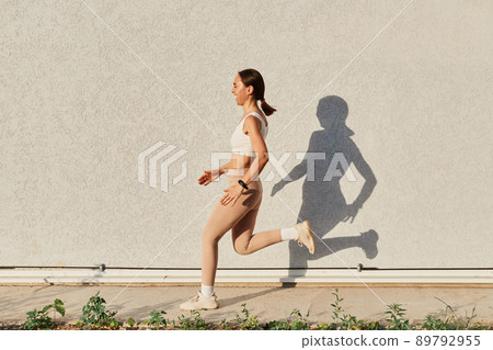 Side view full length portrait of laughing female wearing white top and beige leggins running outside, dark haired woman with ponytail doing with perfect body. 89792955