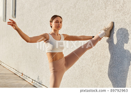 Portrait of beautiful lady wearing white top and beige leggins stretching leg while leaning on gray wall outdoor, spreading hand aside, looking in distance. 89792963