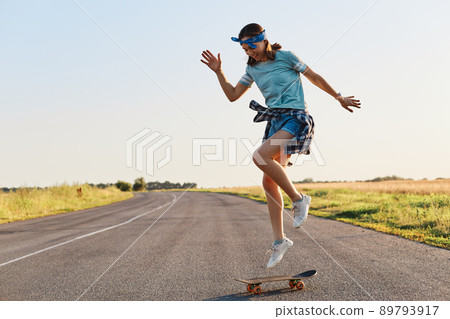 Young sporty woman riding on the longboard on asphalt road, doing tricks on a skateboard, female wearing t shirt, shorts, hair band, having fun alone outdoor. 89793917