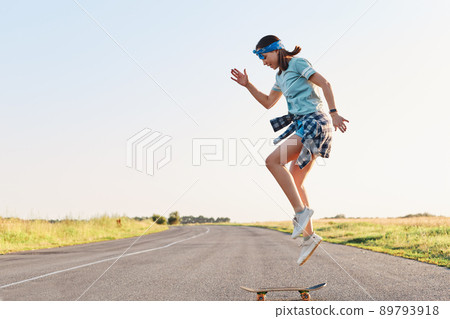 Sporty female wearing t shirt and short doing tricks on a skateboard on street on asphalt road, jumping in the air, enjoying skateboarding alone in sunset in summertime. 89793918