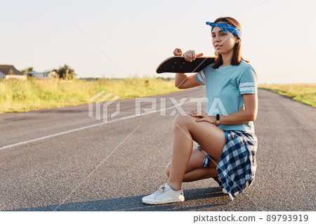 Young adult attractive girl with dark hair posing with a longboard over shoulders while sitting on the road in summer, looking away with pensive expression. Young adult attractive girl with dark hair posing with a longboard over shoulders while sitting on the road in summer, looking away with pensive expression. 89793919