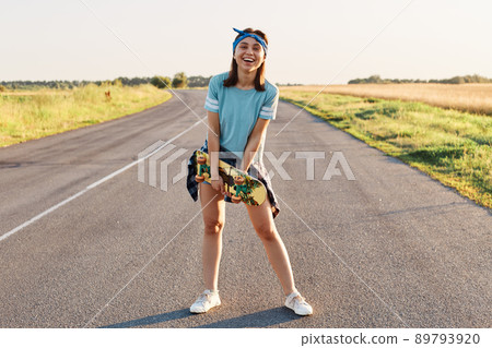Full length portrait of happy laughing female wearing casual t shirt, short and hair band, holding skateboard in hands and looking at camera, healthy lifestyle. Full length portrait of happy laughing female wearing casual t shirt, short and hair band, holding skateboard in hands and looking at camera, healthy lifestyle. 89793920