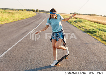 Outdoor shot of sporty woman in casual clothing having fun on asphalt road with skateboard, likes to spend time in active way, healthy lifestyle, happy pastime. 89793921