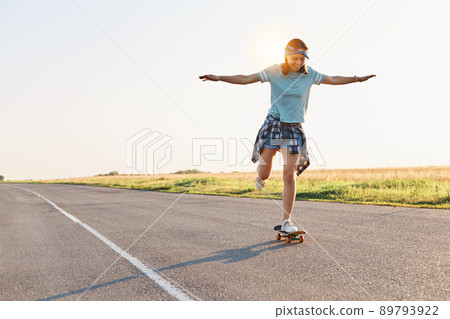 Full length photo of slim attractive happy female with positive emotions and toothy smile, riding skateboard in street, spending summertime in active way. 89793922