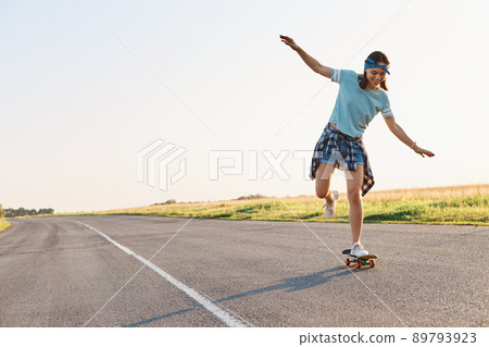 Full length portrait of happy young adult female skateboarding outdoor on asphalt road alone, raised arms, wearing casual style clothing, expressing positive emotions. Full length portrait of happy young adult female skateboarding outdoor on asphalt road alone, raised arms, wearing casual style clothing, expressing positive emotions. 89793923