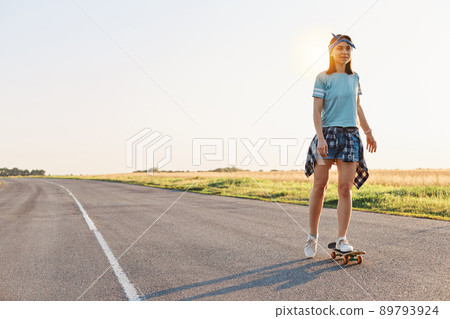 Beautiful female wearing t shirt, short and hair band skateboarding in the street, looking in distance, spending time alone with pleasure, active and healthy lifestyle. Beautiful female wearing t shirt, short and hair band skateboarding in the street, looking in distance, spending time alone with pleasure, active and healthy lifestyle. 89793924