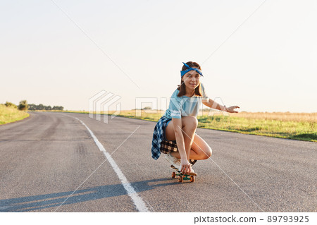 Portrait of sporty woman wearing casual clothing squatting on skateboard, looking in distance with concentrated facial expression, enjoying skateboarding, healthy lifestyle. Portrait of sporty woman wearing casual clothing squatting on skateboard, looking in distance with concentrated facial expression, enjoying skateboarding, healthy lifestyle. 89793925