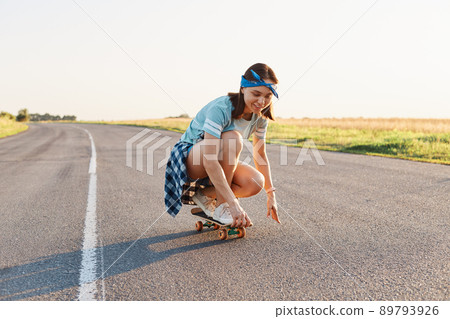 Portrait of happy beautiful dark haired female wearing casual attire and hair band surfing sitting on skateboard, having fun outdoor alone, healthy active lifestyle. Portrait of happy beautiful dark haired female wearing casual attire and hair band surfing sitting on skateboard, having fun outdoor alone, healthy active lifestyle. 89793926