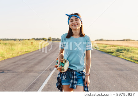 Happy positive female wearing short, blue t-shirt and hair band, standing and looking at camera, holding skateboard in hands, spending time outdoor in summertime. Happy positive female wearing short, blue t-shirt and hair band, standing and looking at camera, holding skateboard in hands, spending time outdoor in summertime. 89793927