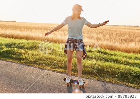 Full length photo of slim beautiful woman wearing casual clothing and hair band skateboarding on asphalt road on sunset, raising arms, enjoying active pastime. Full length photo of slim beautiful woman wearing casual clothing and hair band skateboarding on asphalt road on sunset, raising arms, enjoying active pastime. 89793928