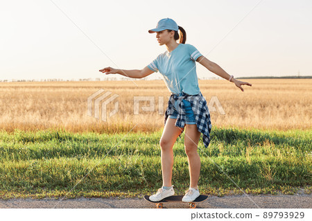 Side view of slim sporty female wearing casual style t shirt and visor cap skateboarding on street on asphalt road, happy active pastime in summer outdoor. Side view of slim sporty female wearing casual style t shirt and visor cap skateboarding on street on asphalt road, happy active pastime in summer outdoor. 89793929