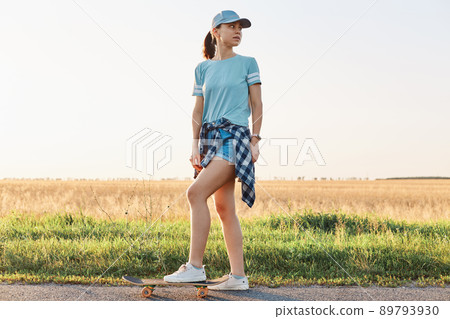 Full length portrait of slim sporty woman wearing t shirt and visor cap, standing with leg on skateboard and looking away, spending her leisure time in active way. Full length portrait of slim sporty woman wearing t shirt and visor cap, standing with leg on skateboard and looking away, spending her leisure time in active way. 89793930
