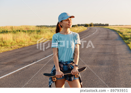 Beautiful female wearing blue t shirt and visor cap standing with skateboard in hands and looking away with toothy smile and positive emotions, active lifestyle. Beautiful female wearing blue t shirt and visor cap standing with skateboard in hands and looking away with toothy smile and positive emotions, active lifestyle. 89793931
