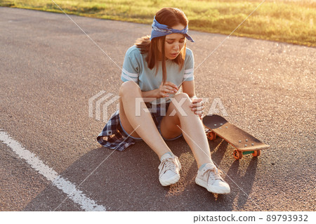 Young adult dark haired woman sitting on asphalt road after falling down from skateboard, injured her knee, feeling pain, looking at her leg with frowning face. 89793932