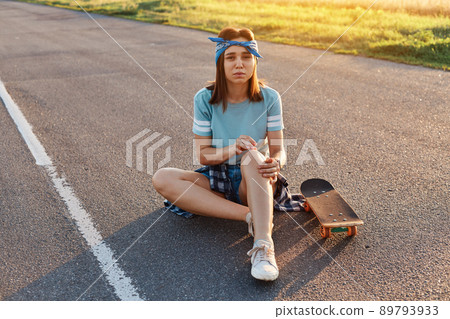 Young adult brunette woman sitting on asphalt road having trauma after falling down from the skate, having pain knee, looking at camera with frowning face. 89793933