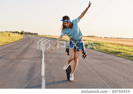 Full length portrait of slim sporty woman doing tricks on a skateboard, spending active time alone, outdoor on street, raised arms, looking down with excited facial expression. Full length portrait of slim sporty woman doing tricks on a skateboard, spending active time alone, outdoor on street, raised arms, looking down with excited facial expression. 89793934