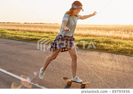Side view of attractive slim sporty female wearing casual attire and hair band skateboarding outdoor alone in sunset, being happy to spend time in active way, summertime. Side view of attractive slim sporty female wearing casual attire and hair band skateboarding outdoor alone in sunset, being happy to spend time in active way, summertime. 89793936