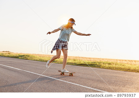 Young attractive female skater riding outdoor on asphalt road, raised her arms, sporty woman wearing casual clothing skateboarding alone in sunset in summer. Young attractive female skater riding outdoor on asphalt road, raised her arms, sporty woman wearing casual clothing skateboarding alone in sunset in summer. 89793937