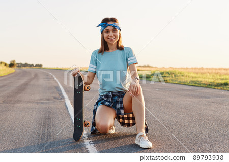 Young adult smiling female with pleasant appearance squatting outdoor on asphalt road and holding skateboard, resting after riding, looking at camera with happy expression. Young adult smiling female with pleasant appearance squatting outdoor on asphalt road and holding skateboard, resting after riding, looking at camera with happy expression. 89793938