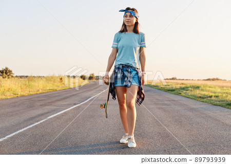 Positive attractive female wearing short, t shirt and hair band holding skateboard in hands and looking away, walking on asphalt road on sunset in summertime. Positive attractive female wearing short, t shirt and hair band holding skateboard in hands and looking away, walking on asphalt road on sunset in summertime. 89793939