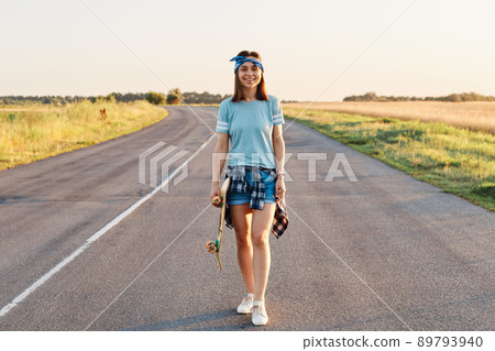 Full length photo of pretty smiling female wearing blue t shirt, short and hair band, standing on asphalt road and holding skateboard in hands, looking at camera with positive emotions. Full length photo of pretty smiling female wearing blue t shirt, short and hair band, standing on asphalt road and holding skateboard in hands, looking at camera with positive emotions. 89793940