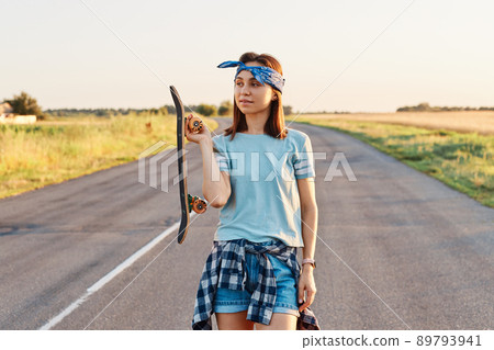 Outdoor shot of beautiful dark haired woman wearing hair band, t shirt and short, holding longboard in hands and looking away with thoughtful expression. Outdoor shot of beautiful dark haired woman wearing hair band, t shirt and short, holding longboard in hands and looking away with thoughtful expression. 89793941