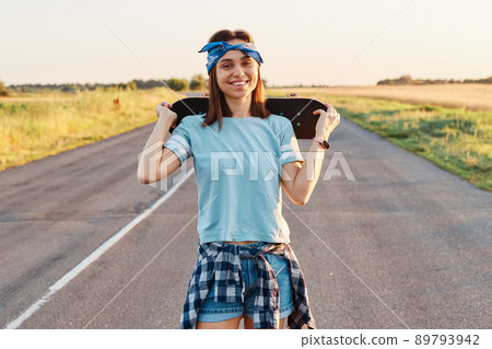 Outdoor portrait of happy dark haired female wearing blue t shirt, short and hair band, looking at camera with toothy smile, holding skateboard over shoulders, expressing happiness. Outdoor portrait of happy dark haired female wearing blue t shirt, short and hair band, looking at camera with toothy smile, holding skateboard over shoulders, expressing happiness. 89793942