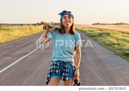 Happy positive female wearing t shirt, short and hair band holding skateboard on her shoulder, looking directly at camera with funny facial expression, grimace, showing tongue out. 89793943