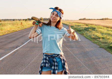 Cheerful girl with stylish hairband posing with a longboard over shoulders while walking on the road, looking at camera with happy emotions and showing thumb up. Cheerful girl with stylish hairband posing with a longboard over shoulders while walking on the road, looking at camera with happy emotions and showing thumb up. 89793944