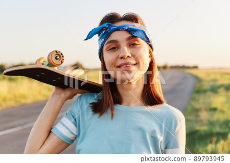 Portrait of beautiful brunette female wearing blue t shirt and hair band, holding skateboard over shoulder and looking at camera, enjoying skateboarding in summertime, active pastime. Portrait of beautiful brunette female wearing blue t shirt and hair band, holding skateboard over shoulder and looking at camera, enjoying skateboarding in summertime, active pastime. 89793945