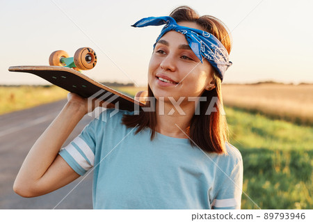 Outdoor shot of smiling brunette woman wearing casual style t shirt and hair band, holding skateboard on shoulders, looking away with smile, expressing happiness. Outdoor shot of smiling brunette woman wearing casual style t shirt and hair band, holding skateboard on shoulders, looking away with smile, expressing happiness. 89793946